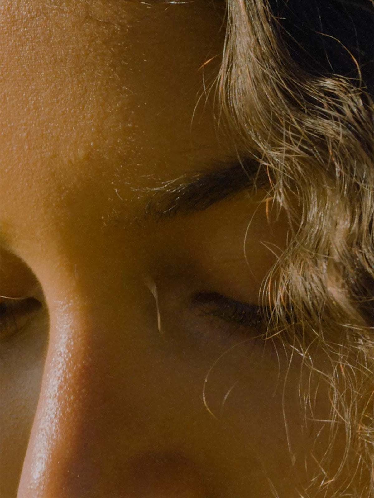 Extreme close-up of a person’s face with eyes lowered, warm sunlight highlighting the skin, eyebrow, nose, and loose curly hair along one side of the image