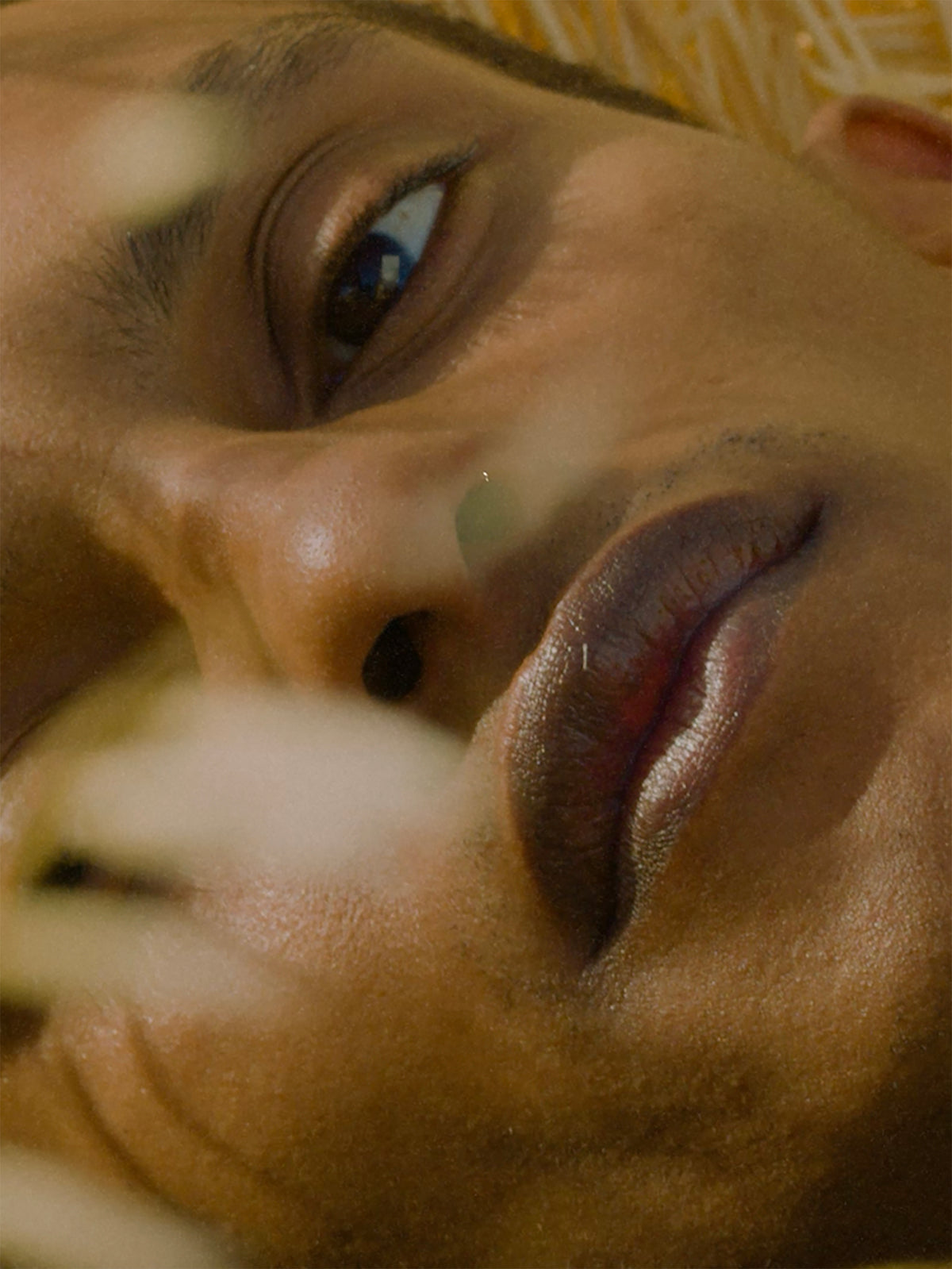 Extreme close-up of a person’s face viewed at an angle, with one eye, textured lips, and soft blurred foliage creating warm highlights across the skin