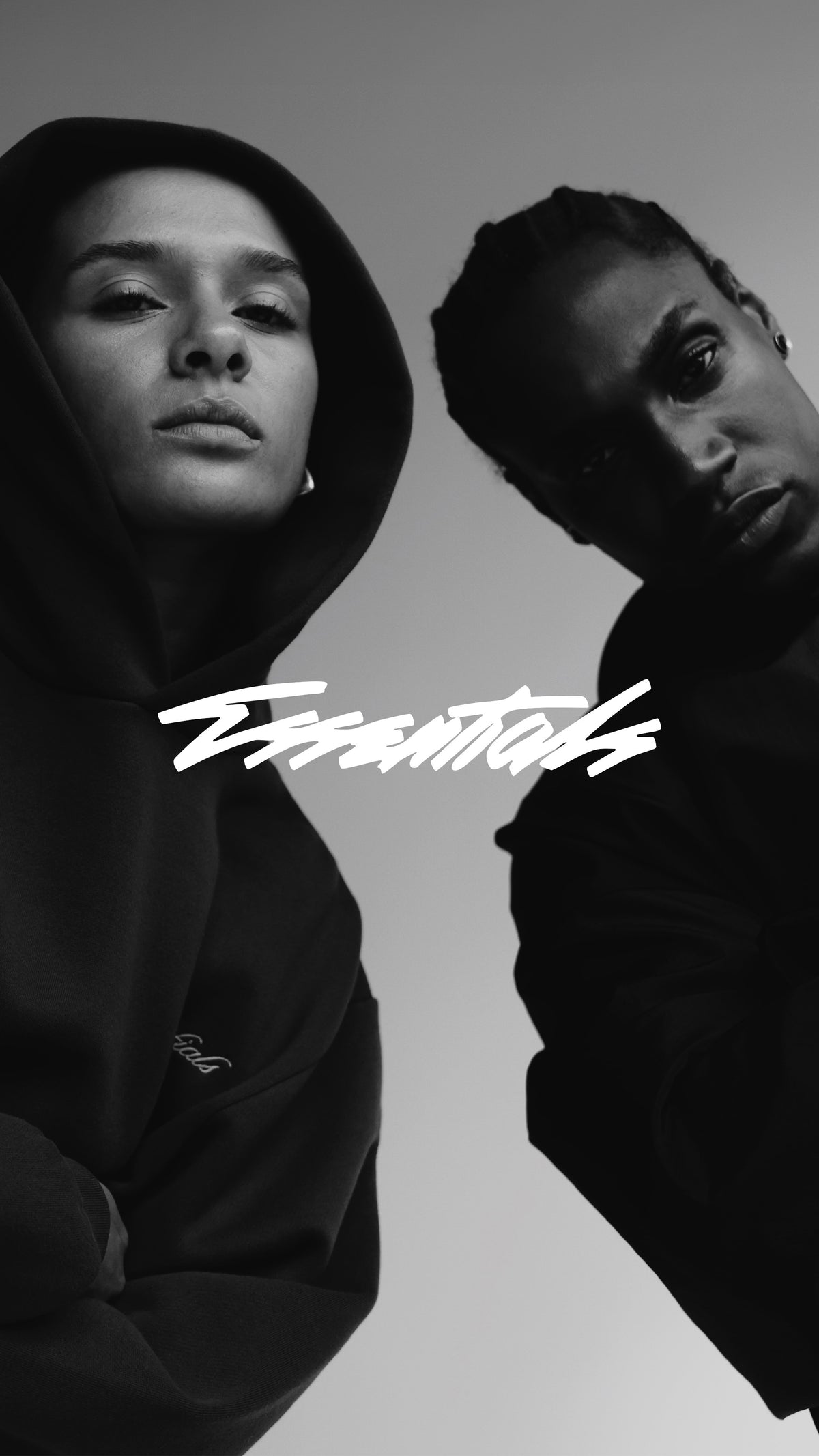 Black and white portrait of two models in dark clothing, one wearing a hood, with a white stylized script logo across the center of the image