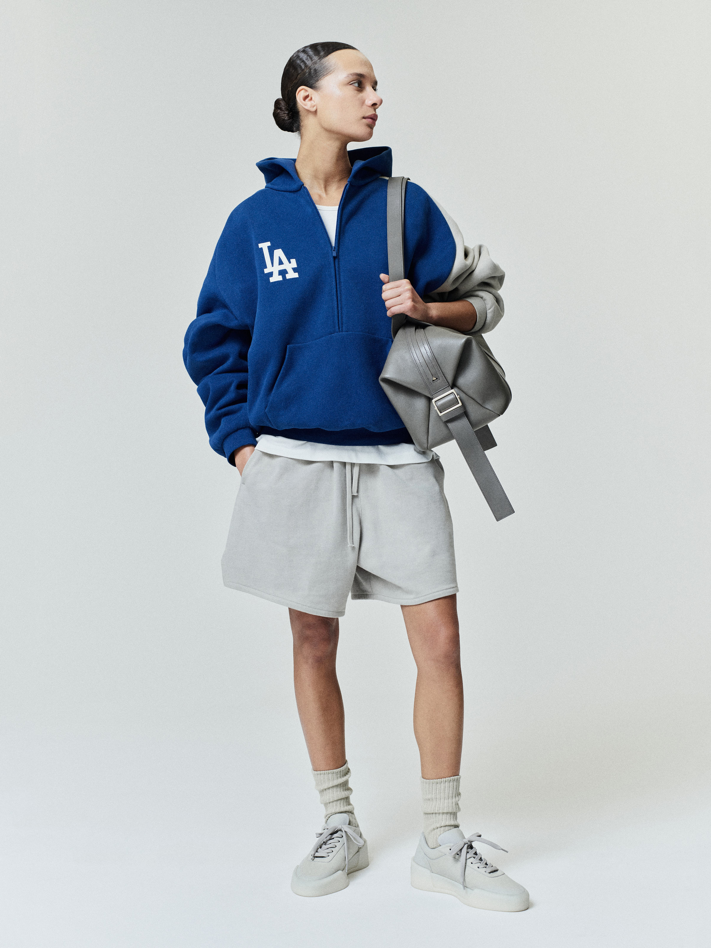 Model wearing a relaxed fit Dodgers Raglan Half Zip Sport Hoodie in deep blue with light gray raglan sleeves, styled with light gray shorts, white sneakers, and a gray shoulder bag.