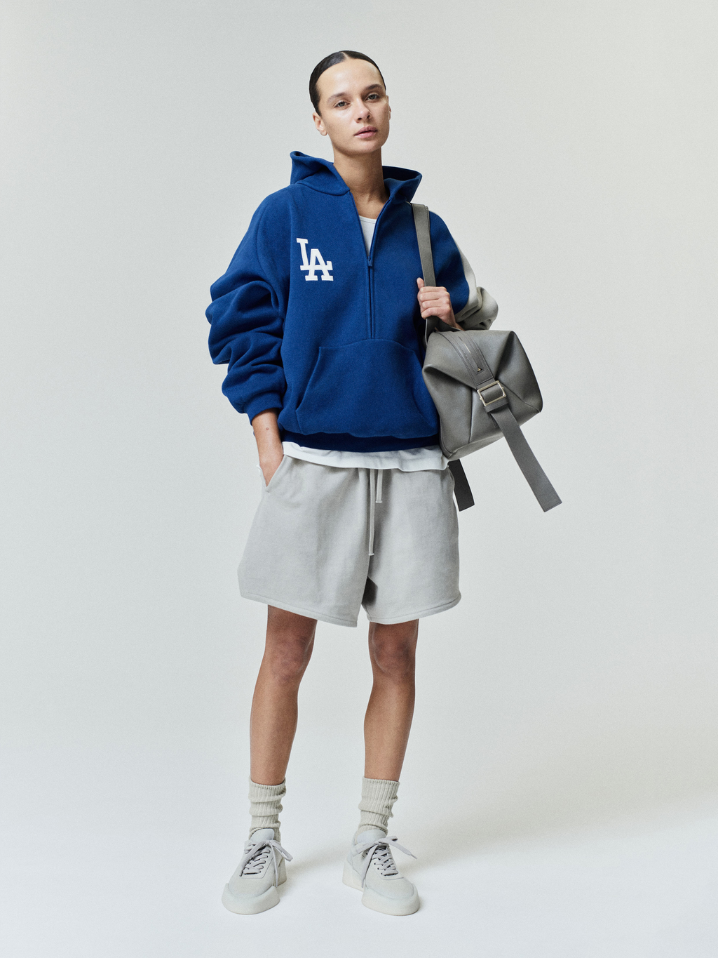 Person wearing a blue hoodie with gray shorts, and carrying a gray backpack on a white background