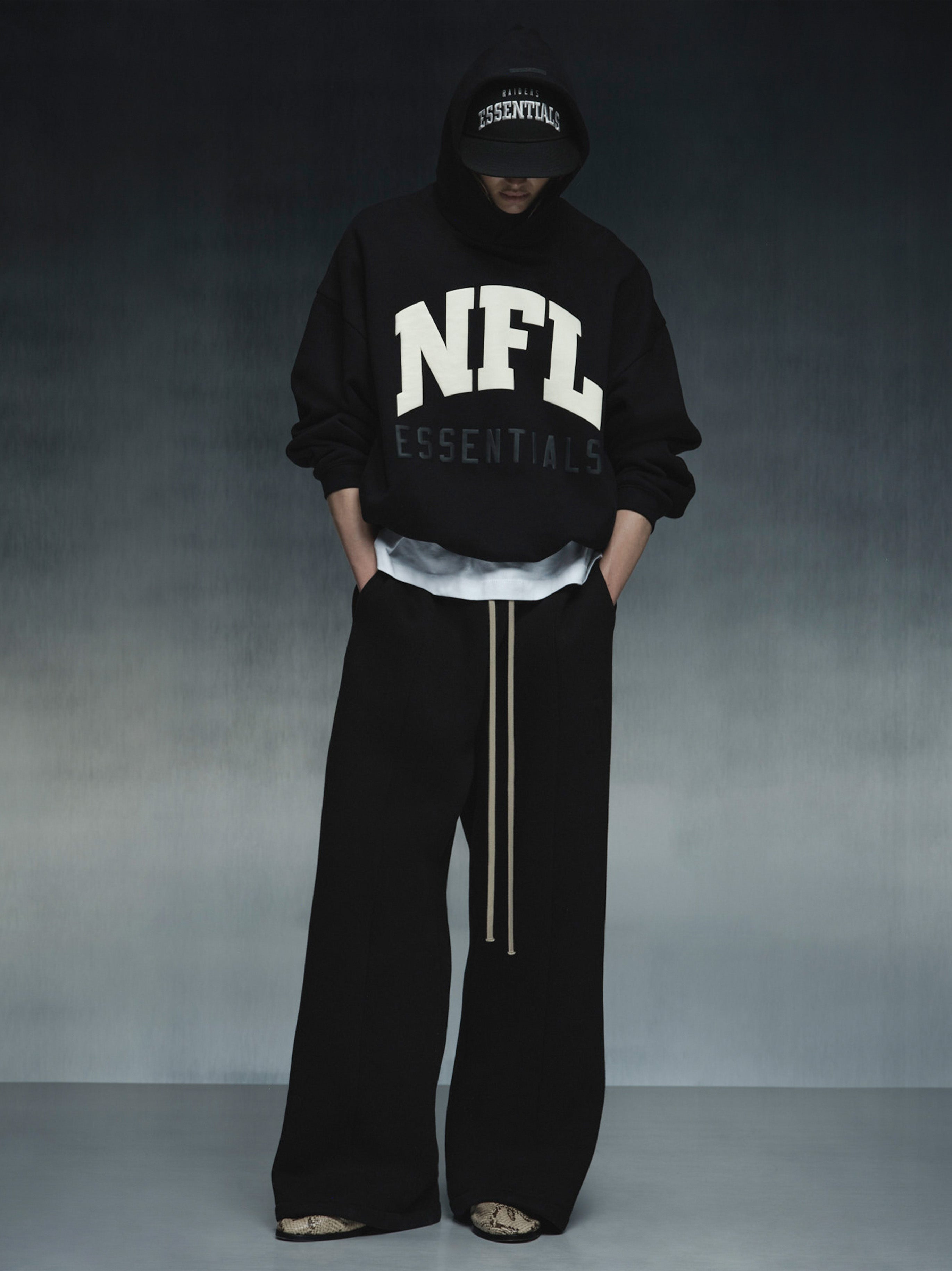 Front view of black NFL Sport Hoodie with large white NFL text and subtle Essentials lettering, styled with black wide pants