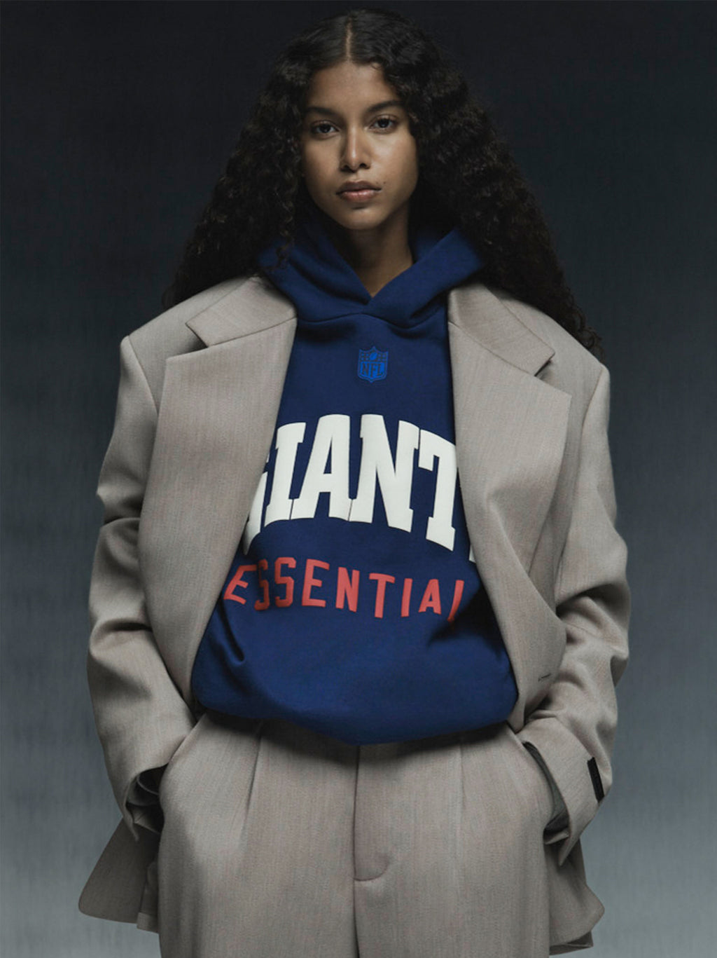 Model wearing blue Giants Sport Hoodie with bold white and red text under beige oversized suit jacket against dark background