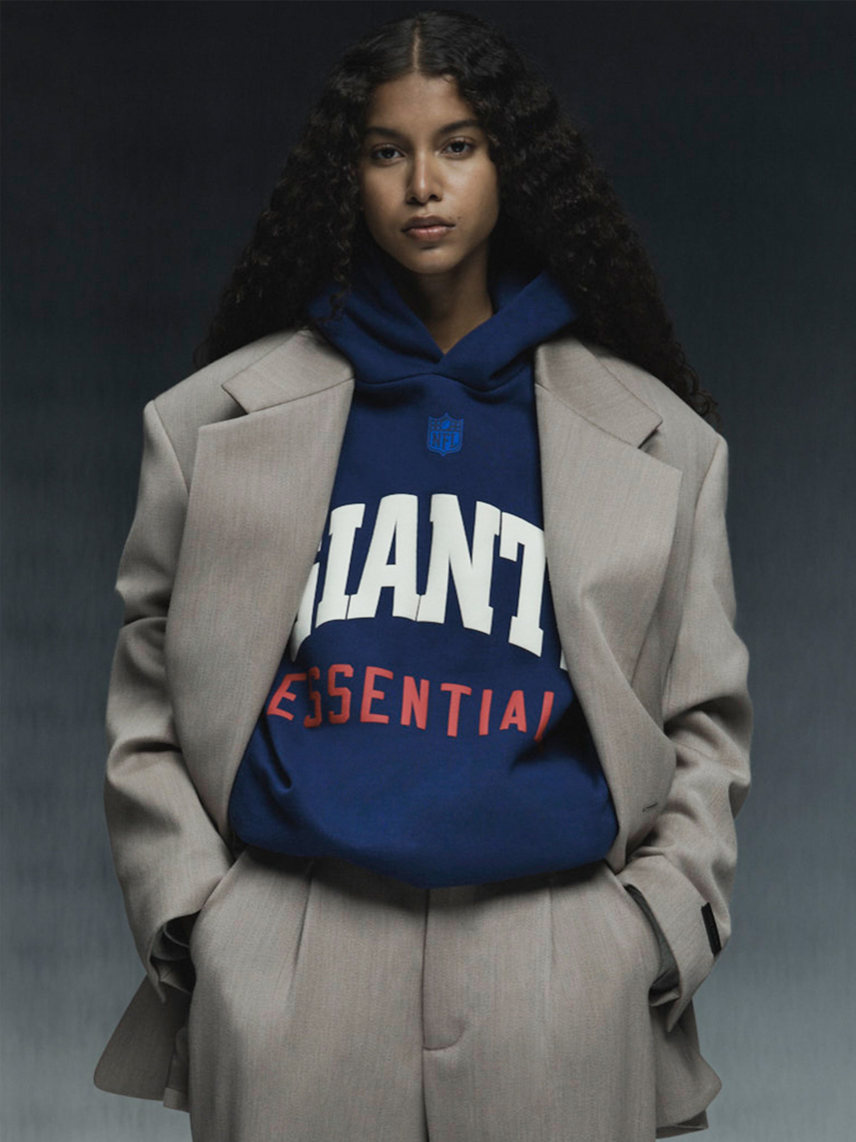 Model wearing blue Giants Sport Hoodie with bold white and red text under beige oversized suit jacket against dark background