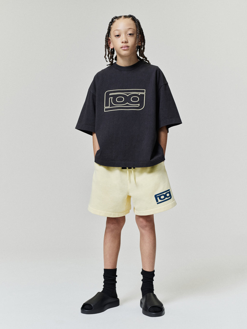 Kids Merch Tee in black with a pale outlined chest graphic styled on a child model with light shorts and black slides as Tops