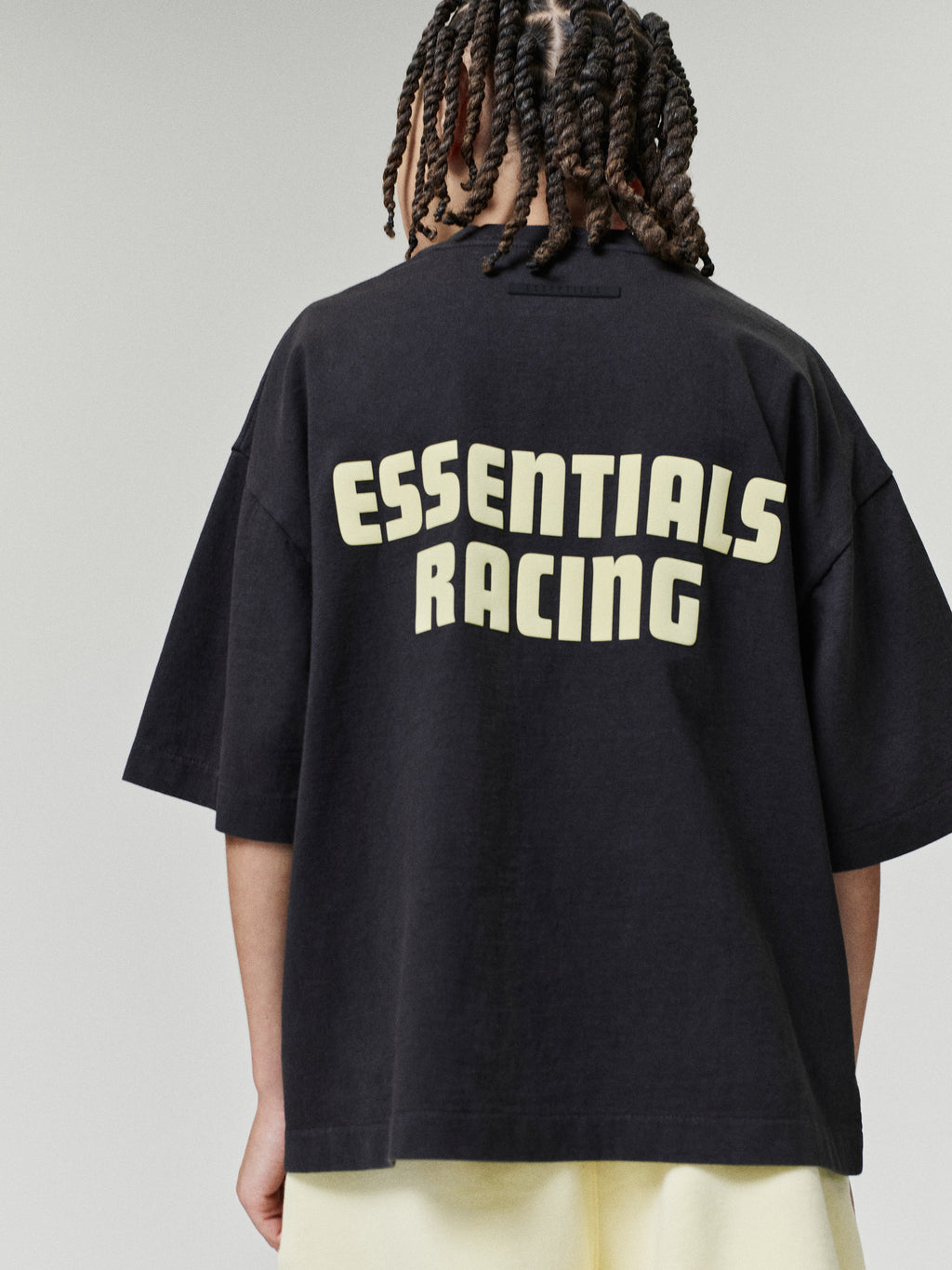 Kids Merch Tee in black shown from behind on a child model with bold cream Essentials Racing lettering across the back paired with light pants as Tops