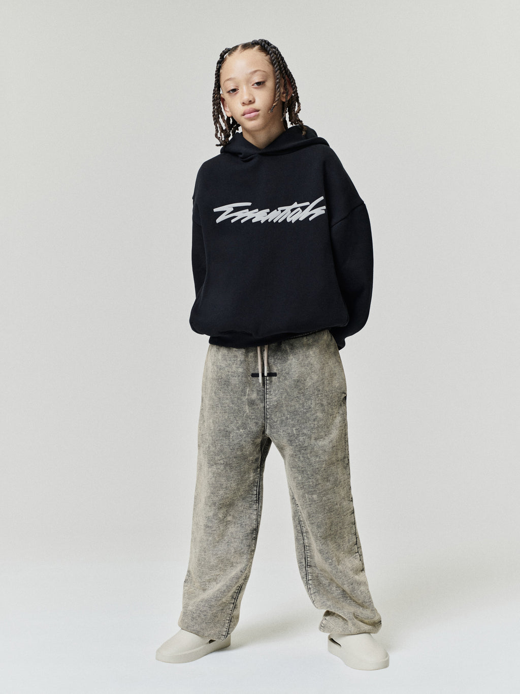 Kids Classic Raglan Hoodie in black with a white script chest graphic styled on a child model with relaxed grey pants and sneakers as Tops
