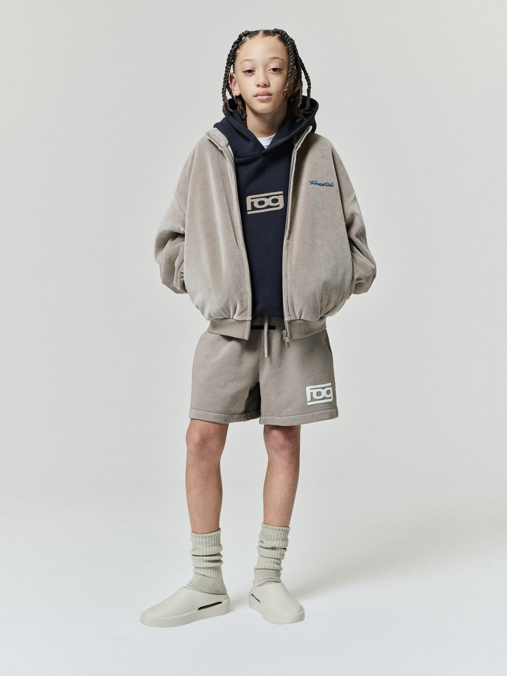 Kids Classic Fit Track Jacket in taupe styled on a child with a zip front elastic cuffs and Fear of God logos layered over a dark hoodie as Outerwear