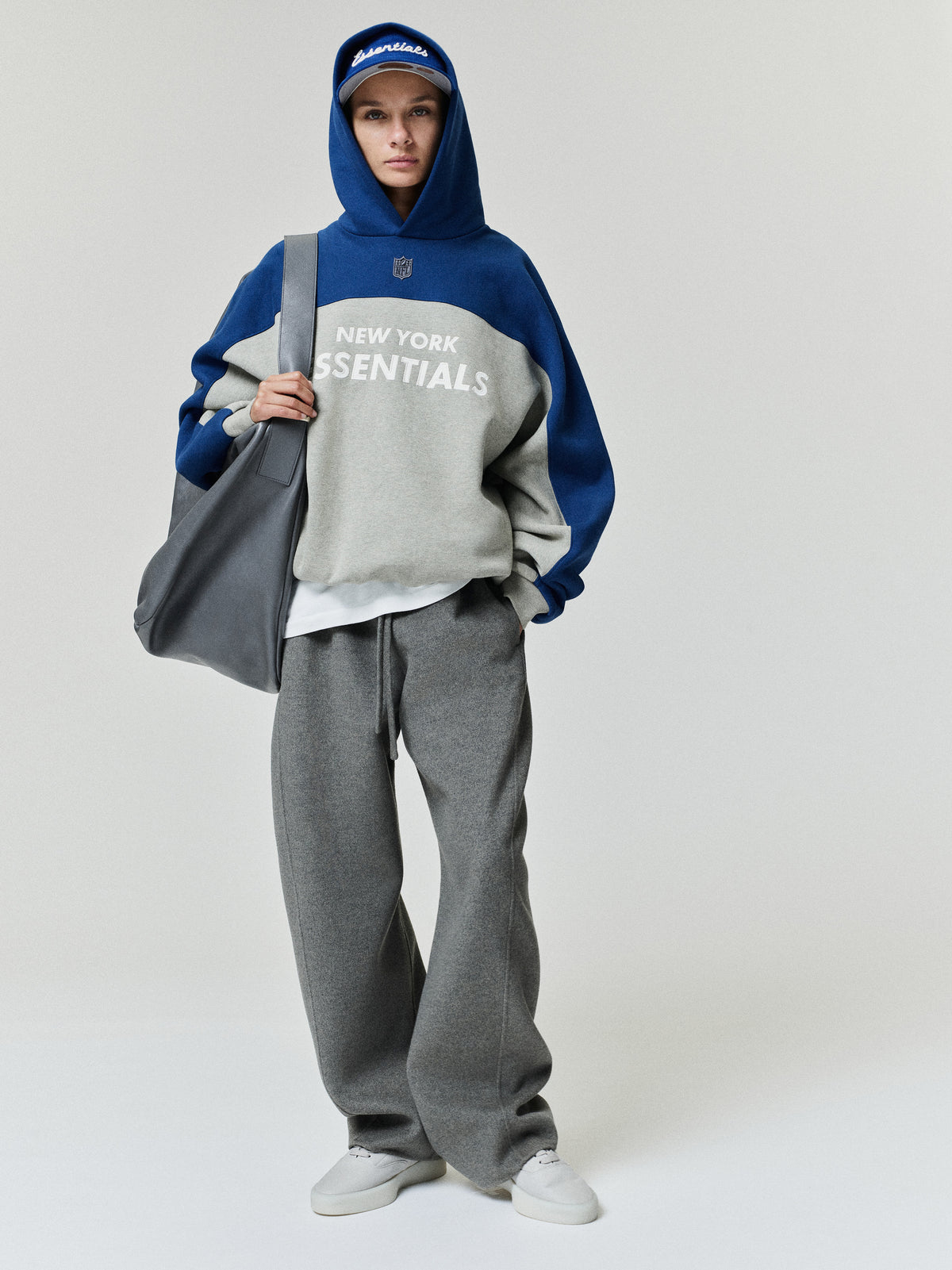 Giants 90s Relaxed Hoodie in blue and gray with New York Essentials lettering worn with gray pants white sneakers and a large tote on a model as a Tops fashion view