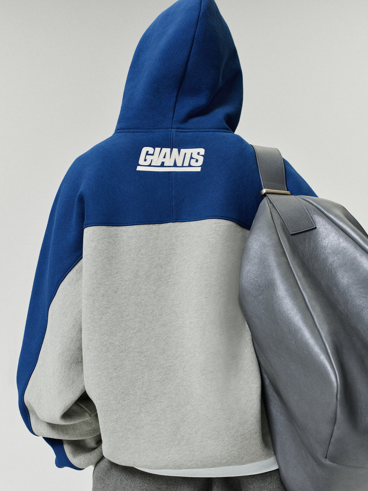 Giants 90s Relaxed Hoodie shown from the back in blue and gray with a white Giants logo and color blocked fleece body styled with a gray backpack as a Tops fashion view