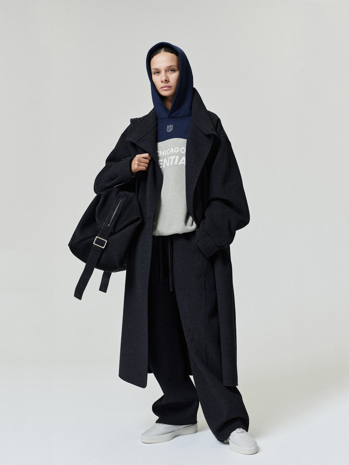 Bears 90s Relaxed Hoodie layered beneath a black overcoat with black trousers and white sneakers on a model carrying a black shoulder bag as a Tops fashion product view
