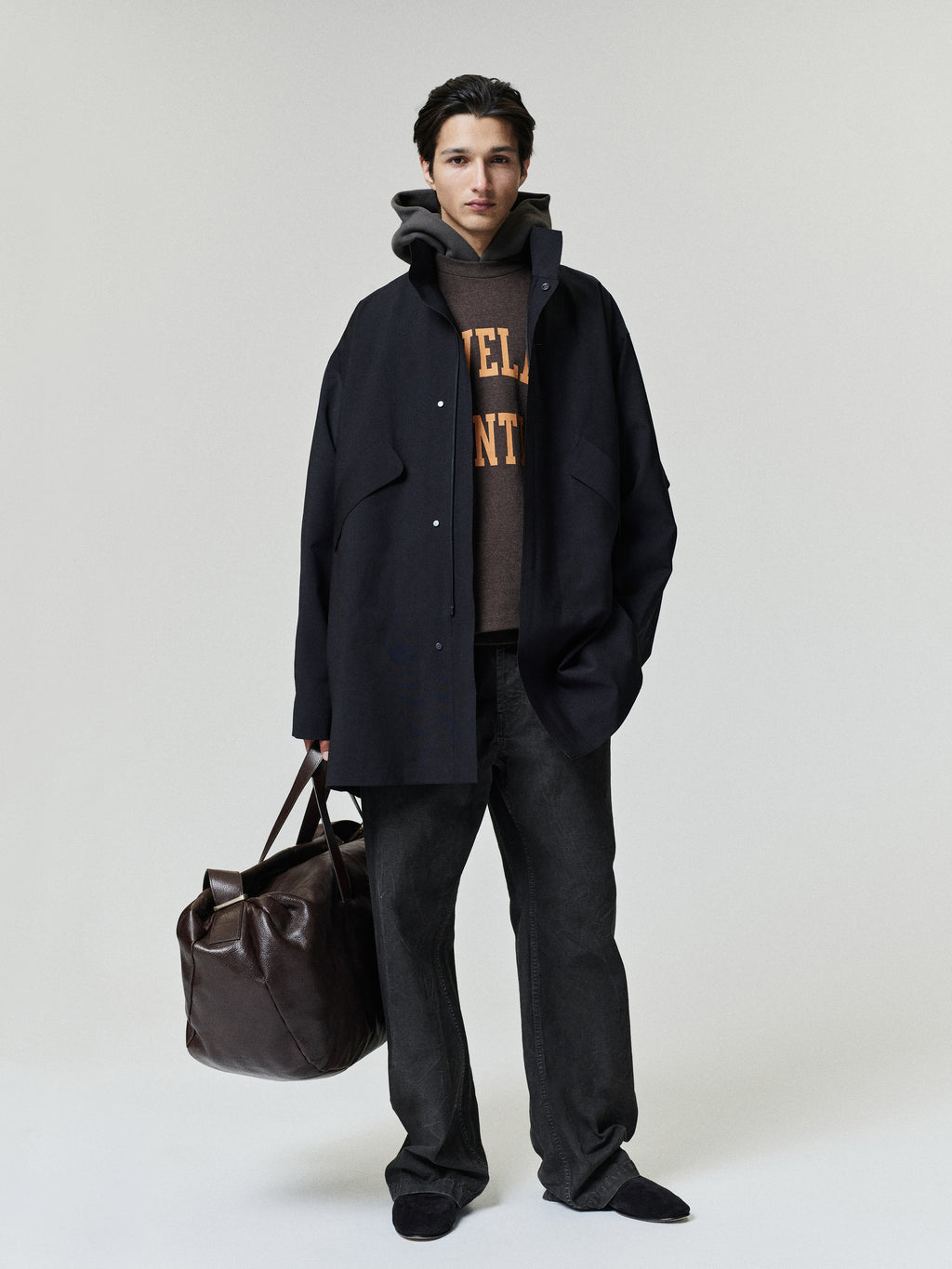 Browns Merch Tee layered under a black overshirt with dark trousers and a brown leather duffle bag in a minimalist Fear of God Tops look