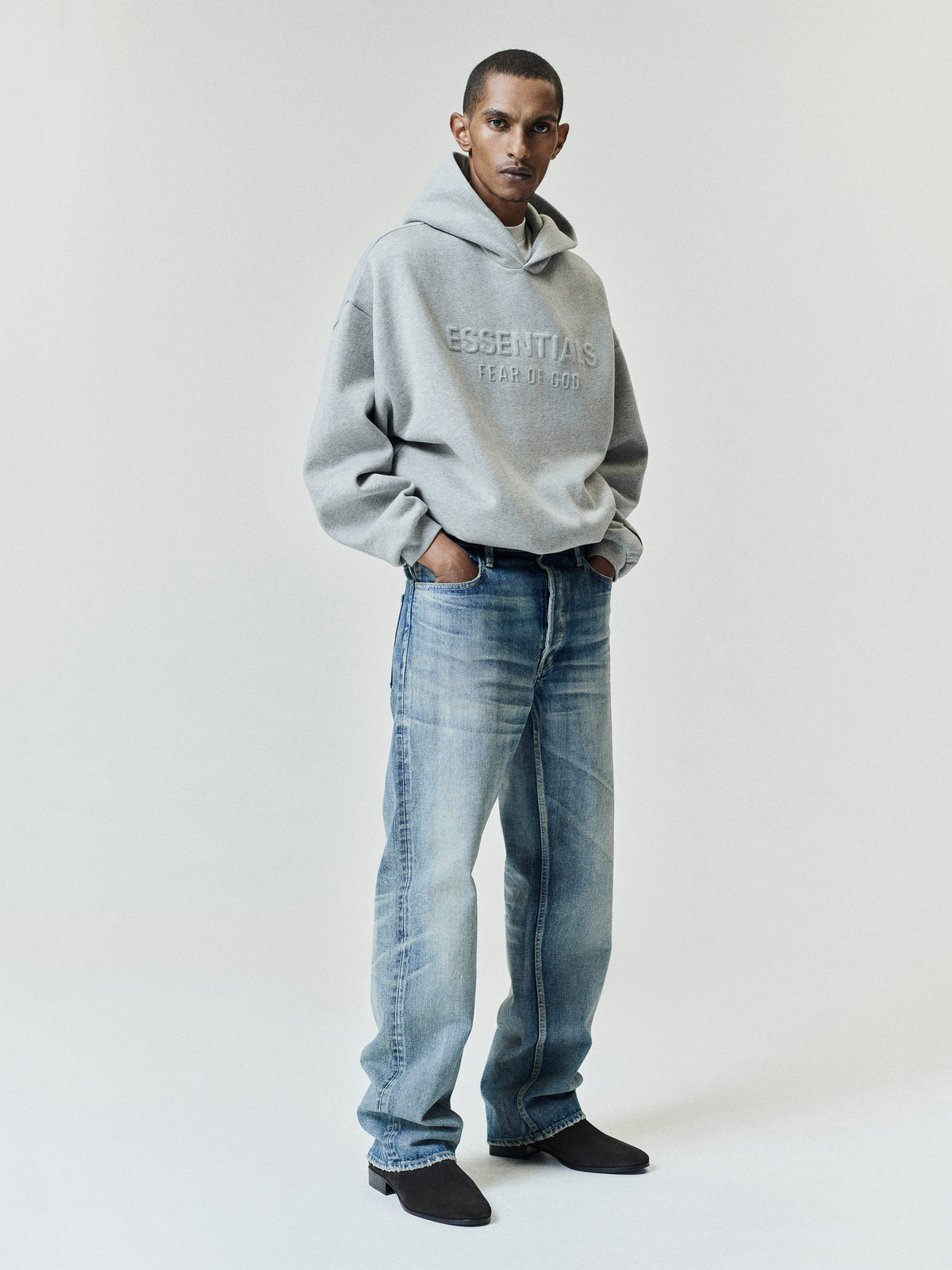 Embossed FW26 90s Relaxed Hoodie in gray with embossed chest lettering worn with relaxed blue jeans and black shoes against a light studio backdrop