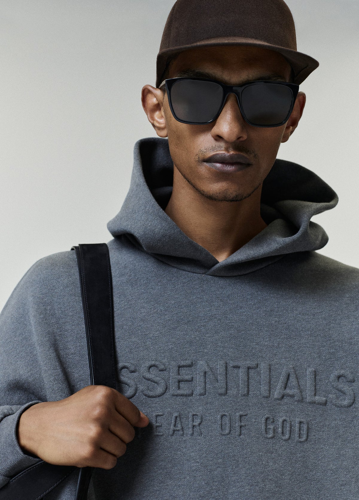 Embossed FW26 90s Relaxed Hoodie in gray with raised chest lettering worn by a model in sunglasses and a cap carrying a black shoulder bag