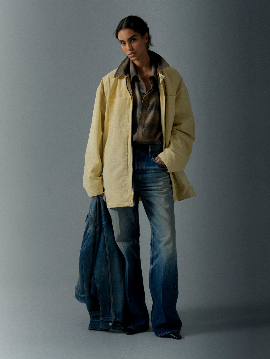 Woman wearing a loose light yellow jacket over a patterned shirt and wide-leg jeans holding a denim jacket against a gray background