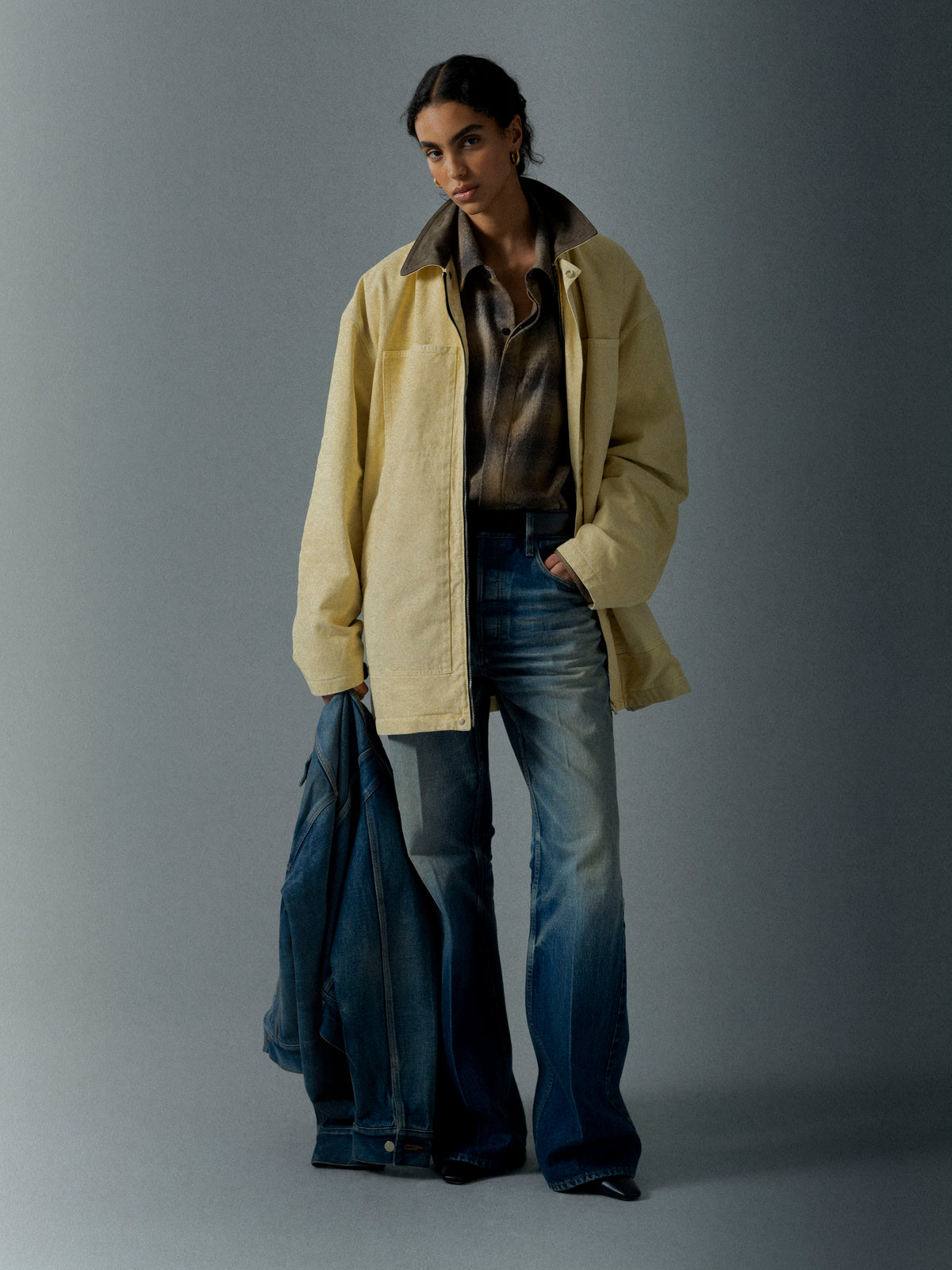 Woman wearing a loose light yellow jacket over a patterned shirt and wide-leg jeans holding a denim jacket against a gray background