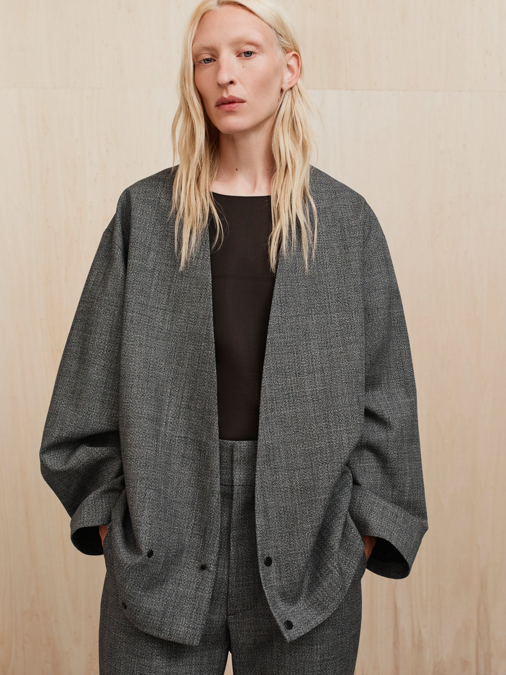 Model wearing oversized gray textured blazer with open front and matching trousers against beige backdrop