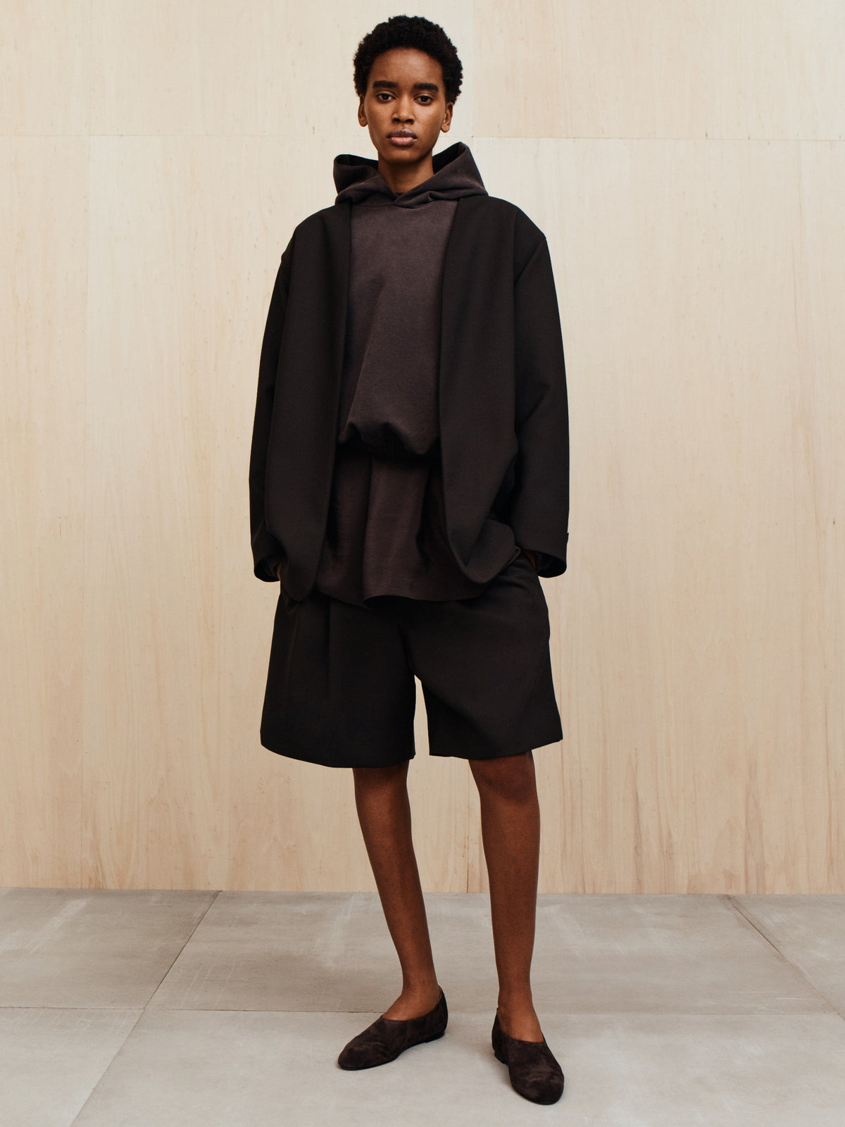 Person wearing a loose black blazer over a dark oversized hoodie and wide black knee-length shorts on a tiled floor against beige wall