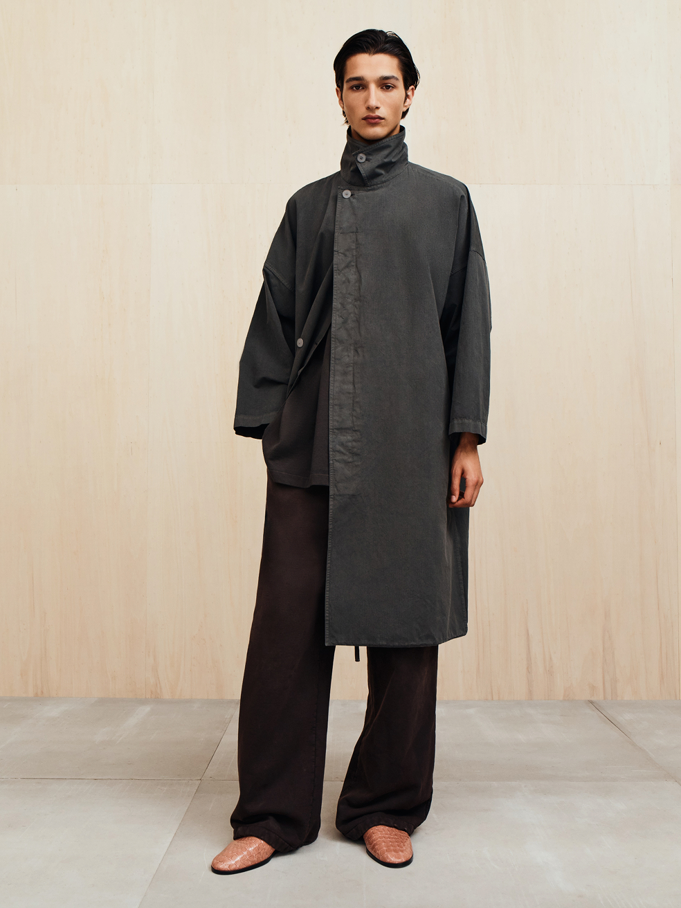 Person wearing a long, dark asymmetrical coat with a high collar, loose dark pants, and brown pointed shoes.