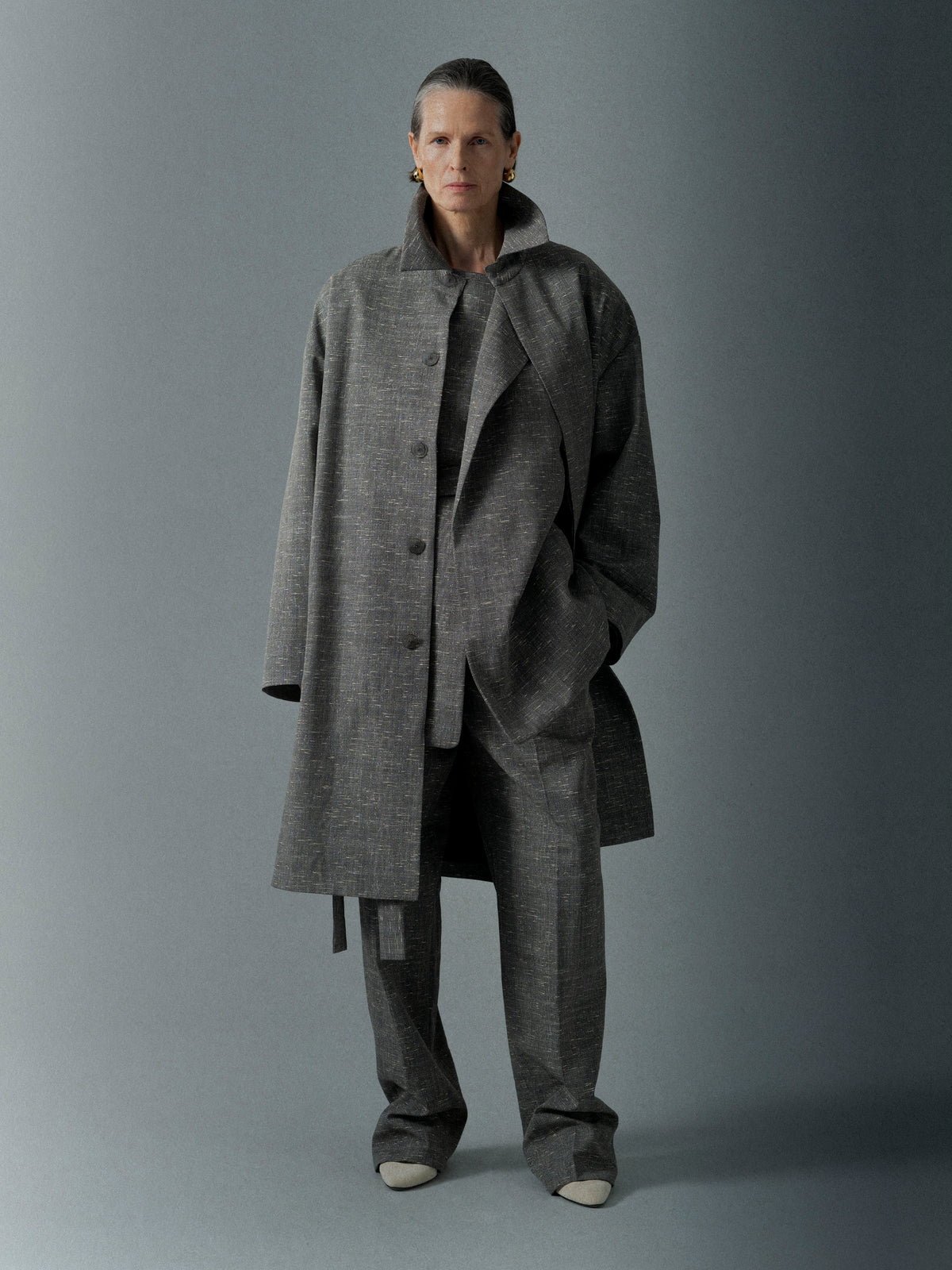 Person with slicked-back hair wearing a loose gray textured coat and matching wide-legged pants against a solid background