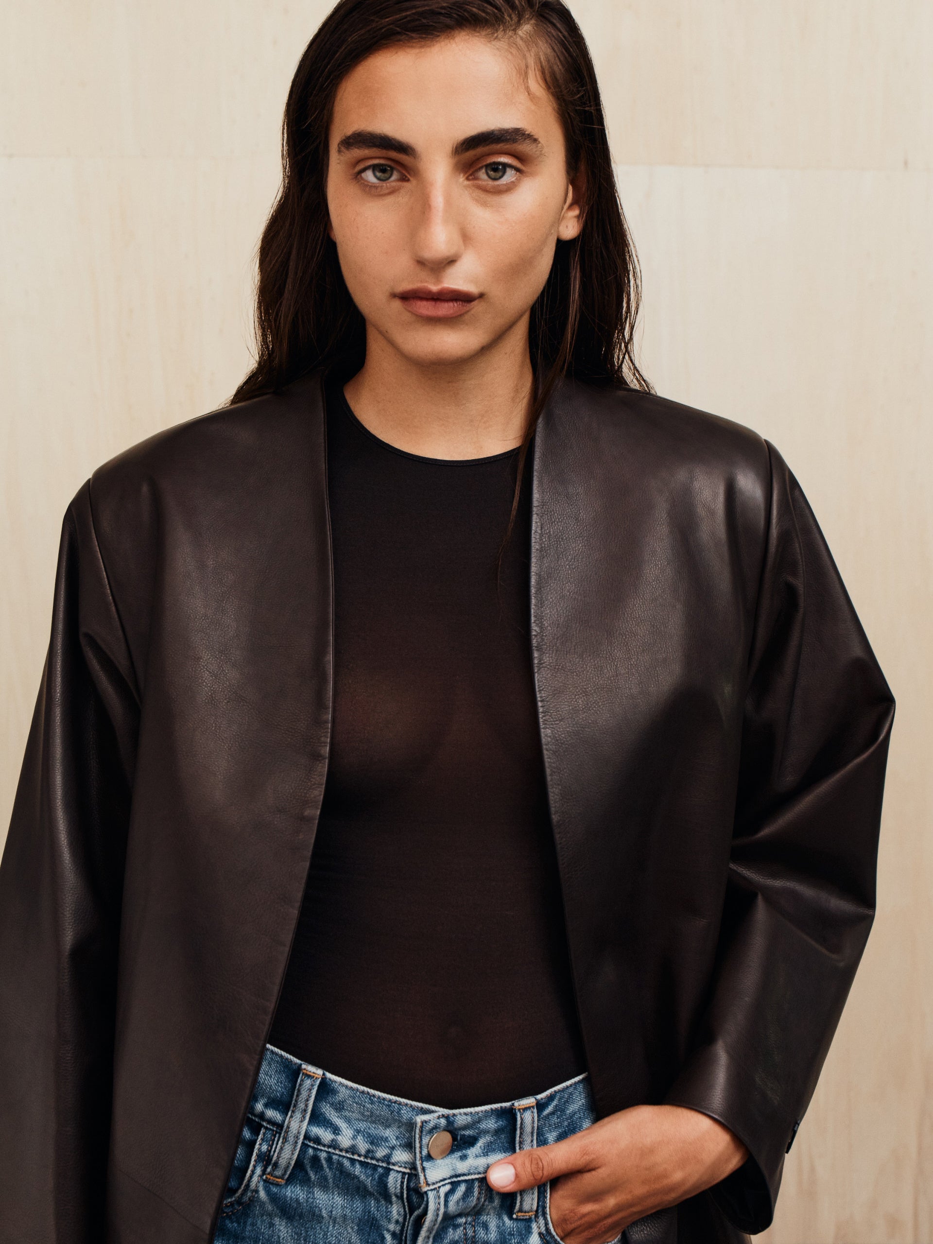 Person with dark hair wearing a loose black leather jacket over a sheer black top and blue jeans standing against a beige wall