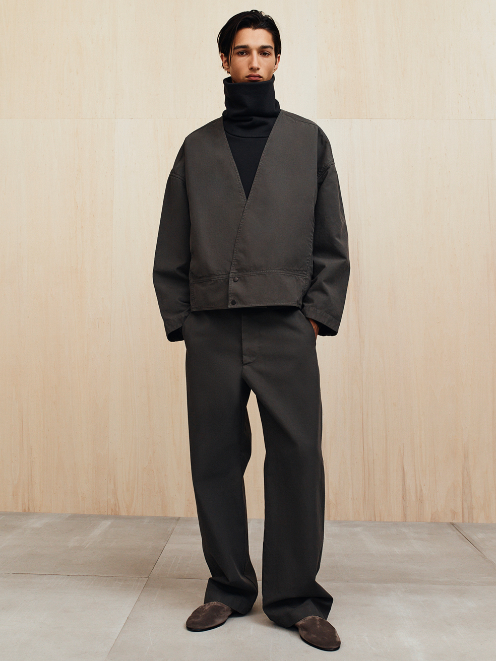 A man wearing a dark oversized jacket, black turtleneck, wide-leg pants, and brown shoes standing against a wooden wall.