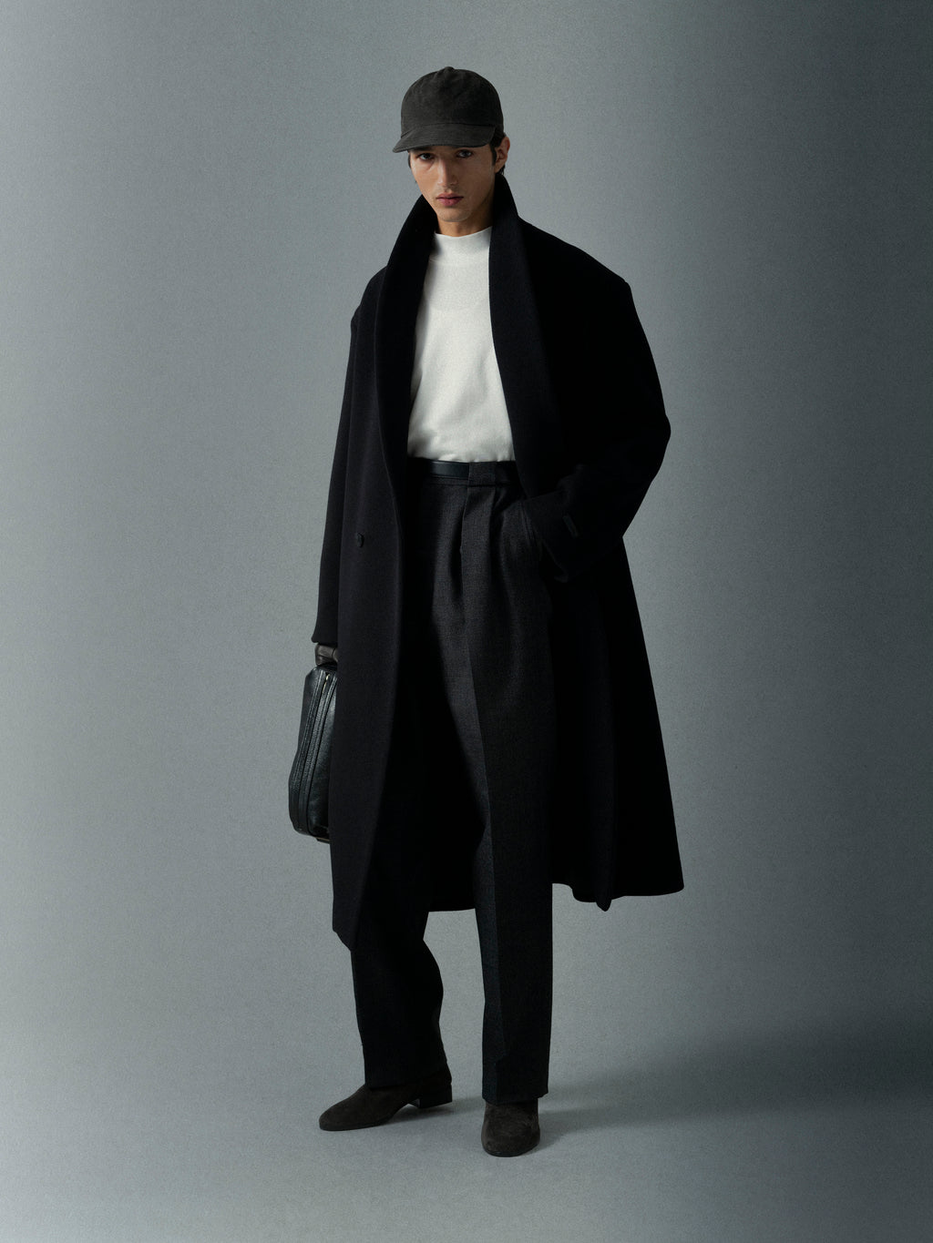Man wearing a long black coat, white shirt, and black pants holding a leather bag against a plain gray background
