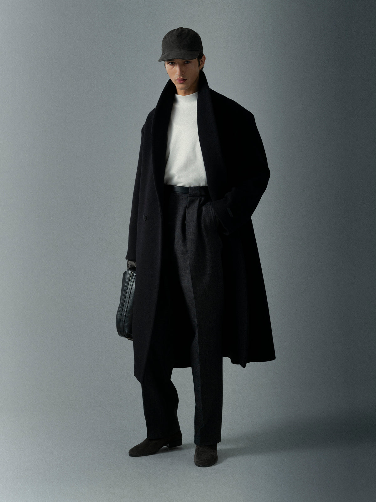 Man wearing a long black coat, white shirt, and black pants holding a leather bag against a plain gray background