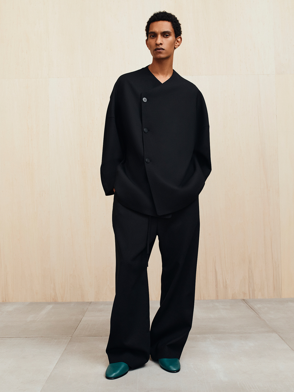 Man wearing loose-fitting black outfit with oversized sleeves and wide-leg pants standing against a beige wall