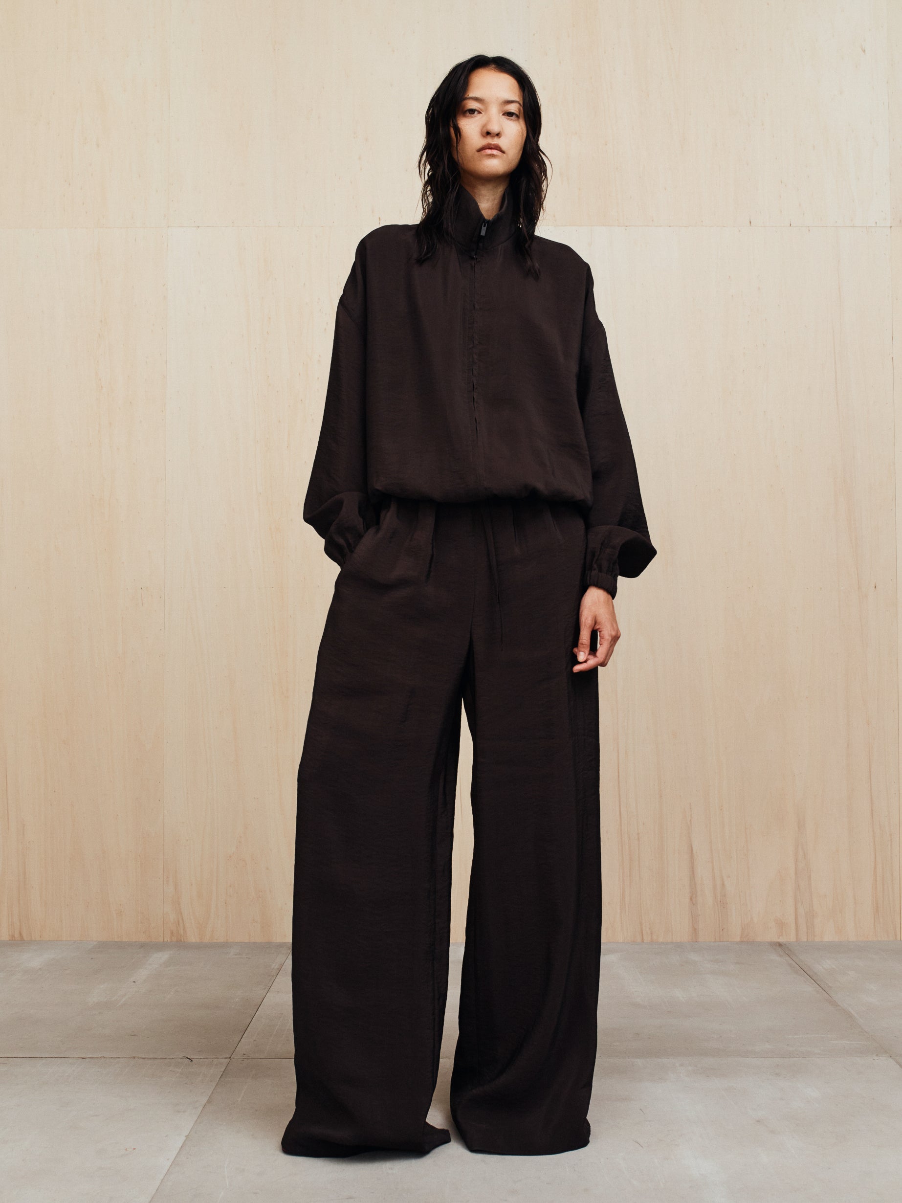 Person with wet dark hair wearing loose black long-sleeve top and wide-leg pants standing against a light wood wall
