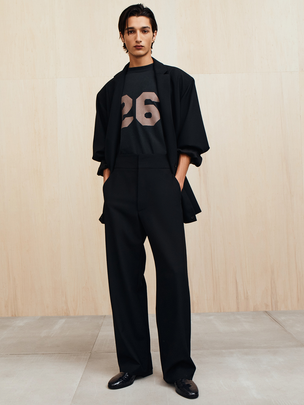 Young man with slicked-back hair wearing a black oversized blazer, black trousers, and a black t-shirt with the number 26 standing against a wood panel background.