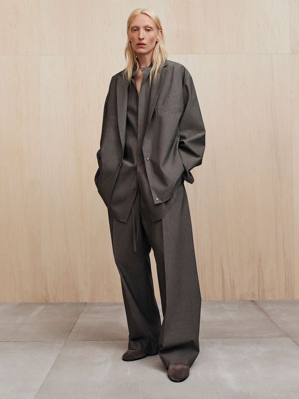 Tall person with long blonde hair wearing an oversized dark gray suit standing against a wooden wall