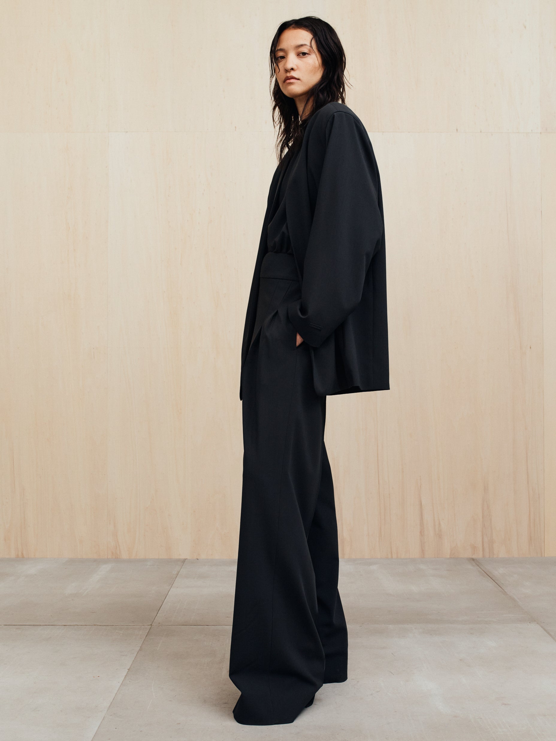 Person with wet dark hair wearing an oversized black blazer and wide-leg black pants standing against a plain wall
