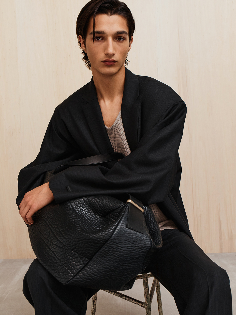 Young man with dark hair sitting on a stool wearing a black blazer and holding a large black textured bag