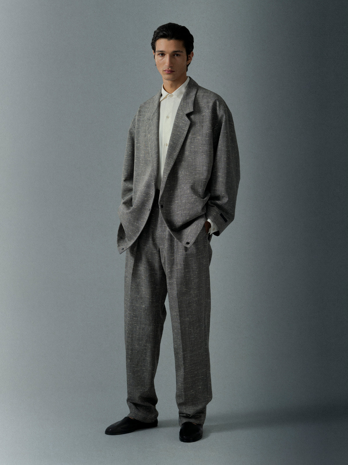 Young man wearing a loose gray checked suit with black shoes and a white shirt standing against a gray background
