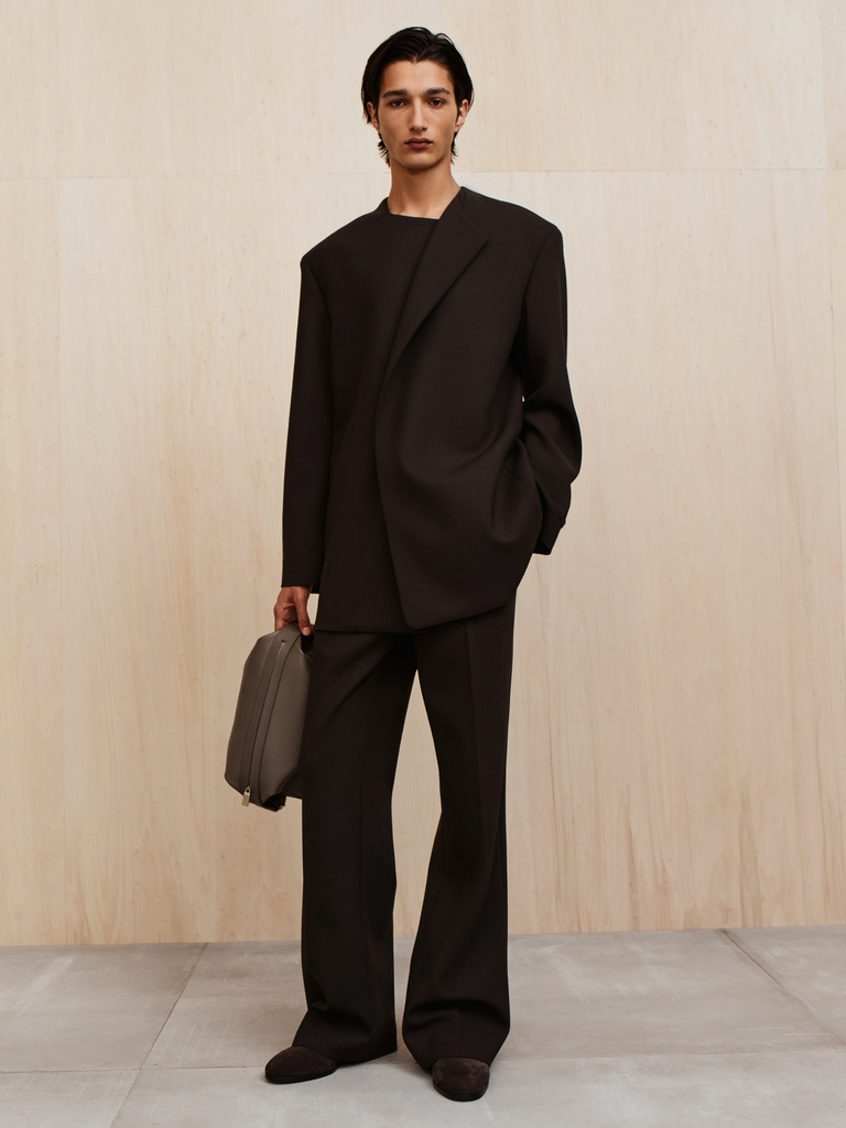 Person with medium-length dark hair wearing an oversized black suit holding a gray bag standing against a light wood wall