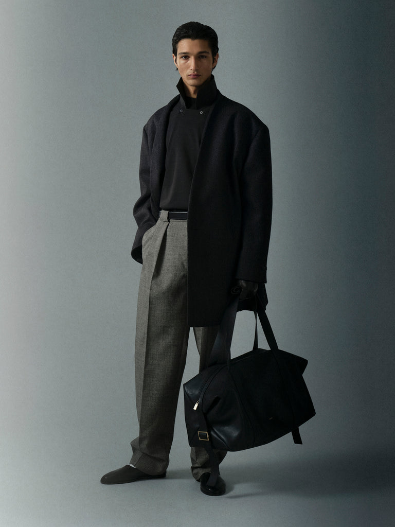 Man wearing dark oversized coat, black turtleneck, gray pleated trousers, black gloves, and holding a large black bag
