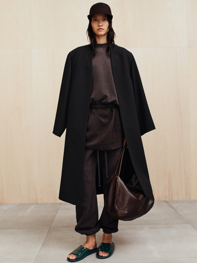 Person wearing oversized black coat over dark casual outfit holding large brown leather bag against light wall