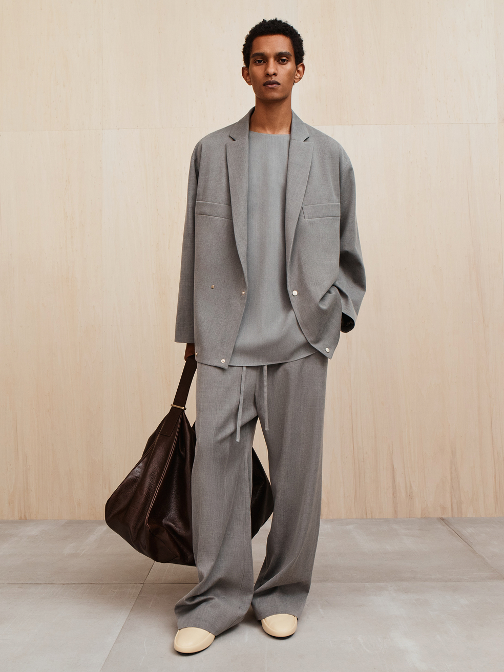 Man wearing loose gray suit with matching top and wide-leg pants carrying large dark brown bag against wooden background