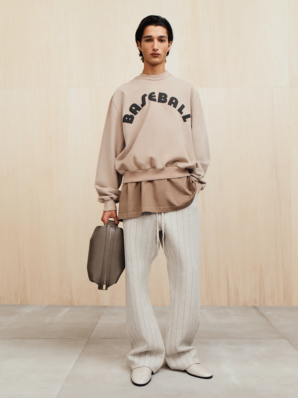 Young person wearing oversized beige sweatshirt with "BASEBALL" text, loose beige pants, and holding a large taupe bag