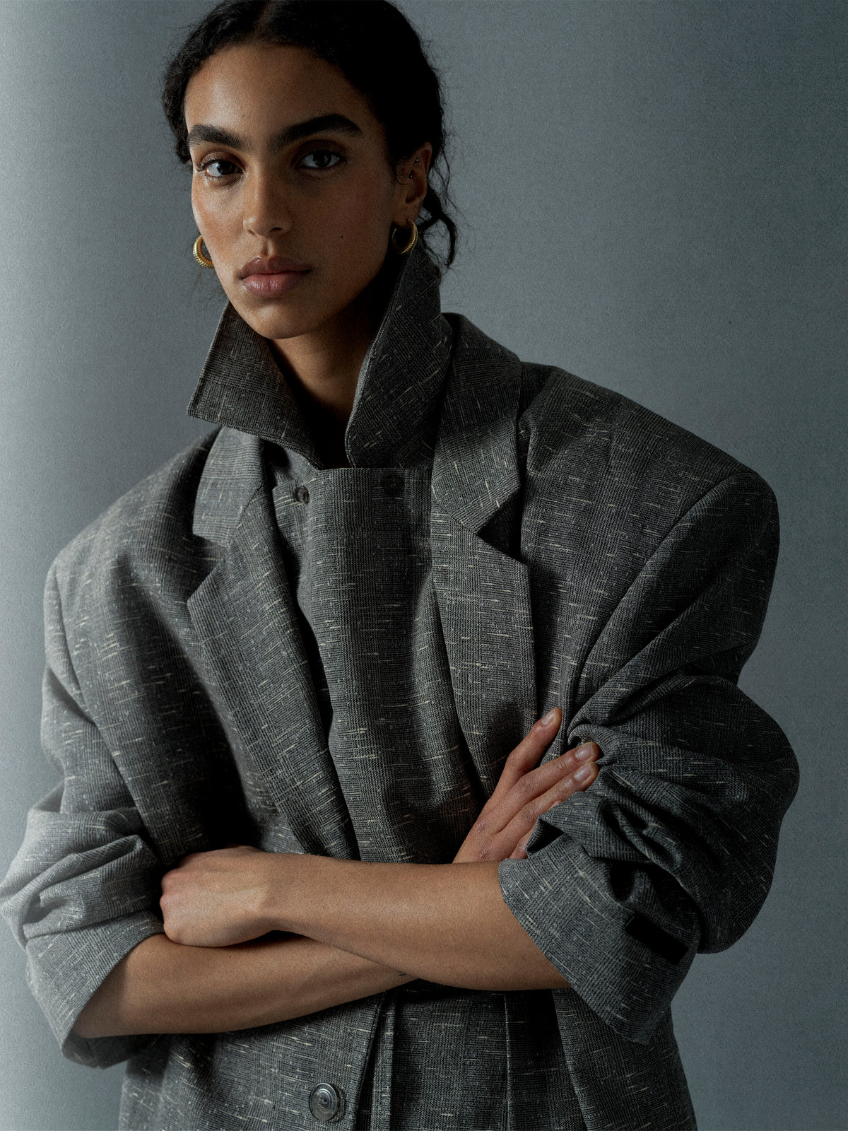 Person with dark hair wearing an oversized gray textured jacket with rolled-up sleeves against a plain backdrop