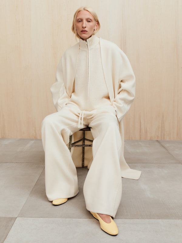 Person with light blonde hair wearing oversized cream knitwear and wide pants seated on a stool against a wooden wall