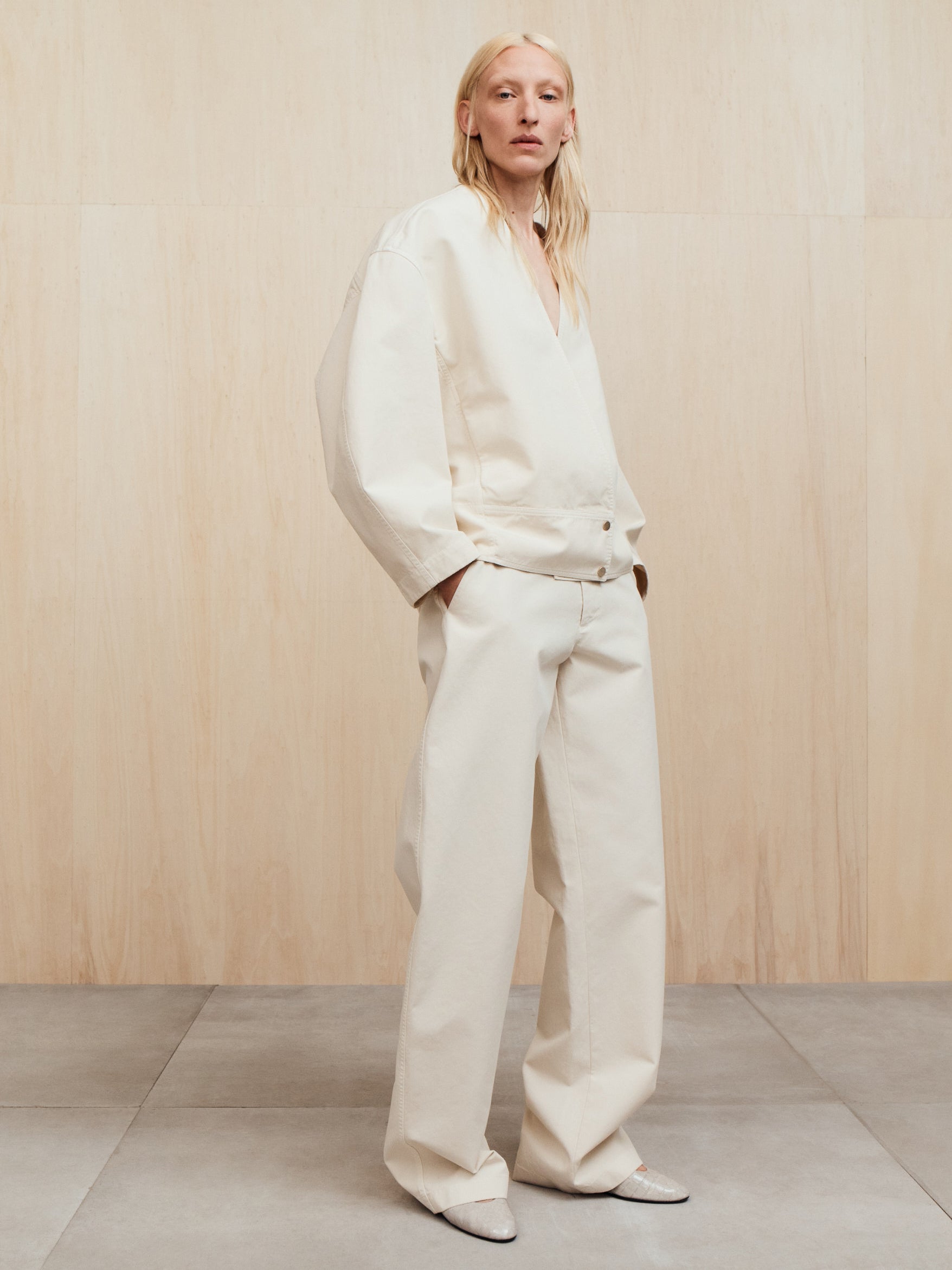 Person with straight light hair wearing loose off-white jacket and wide matching pants standing against a plain wood wall