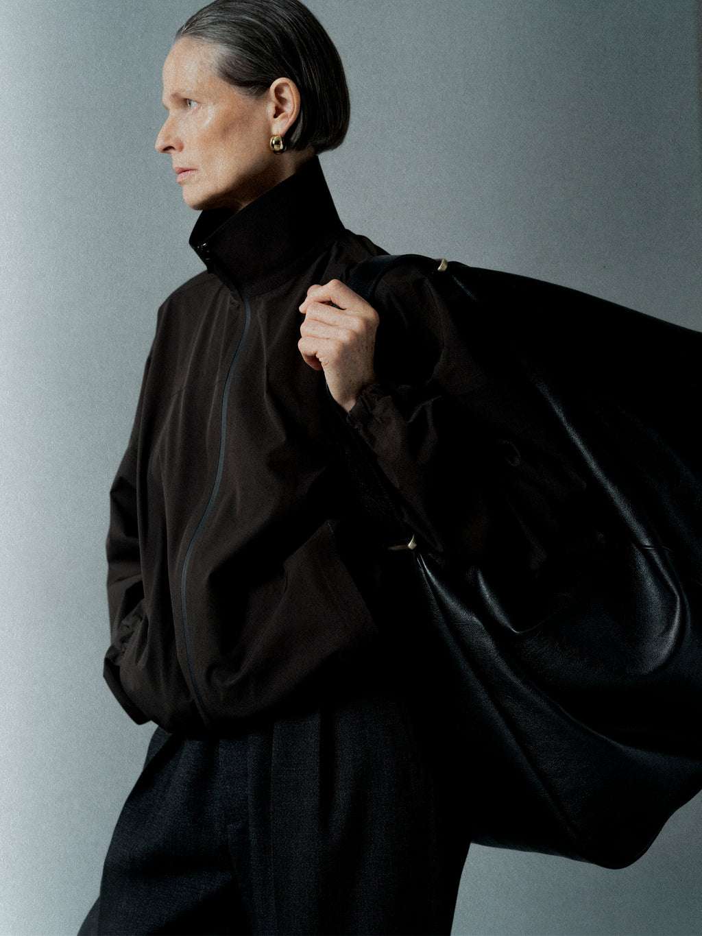 Older woman with sleek gray hair wearing a high-collared black jacket and gold hoop earrings against a plain background