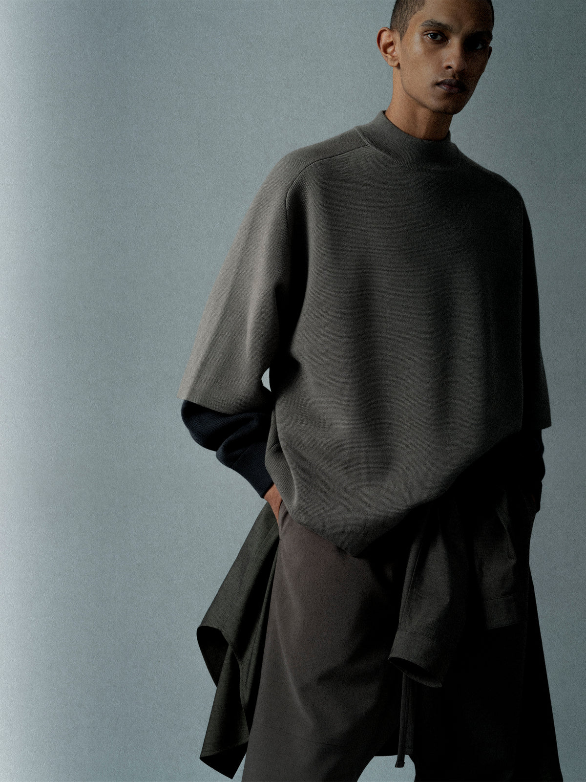 Person wearing a loose dark gray oversized sweater with half sleeves over a black long-sleeve shirt against a plain background