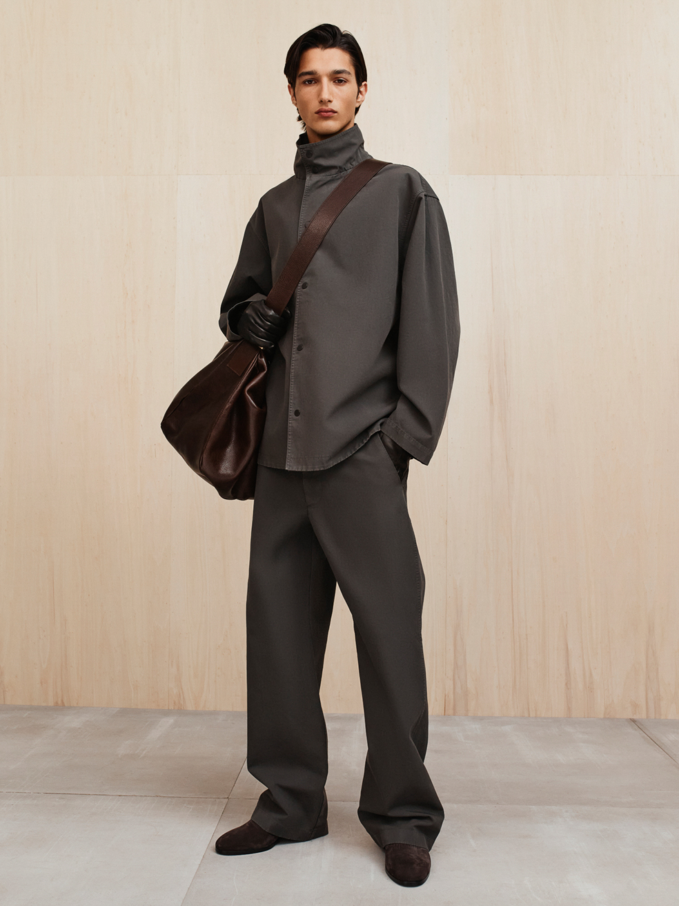 Young man in dark gray loose-fitting outfit with high collar, holding a large brown leather bag against a beige wall background.