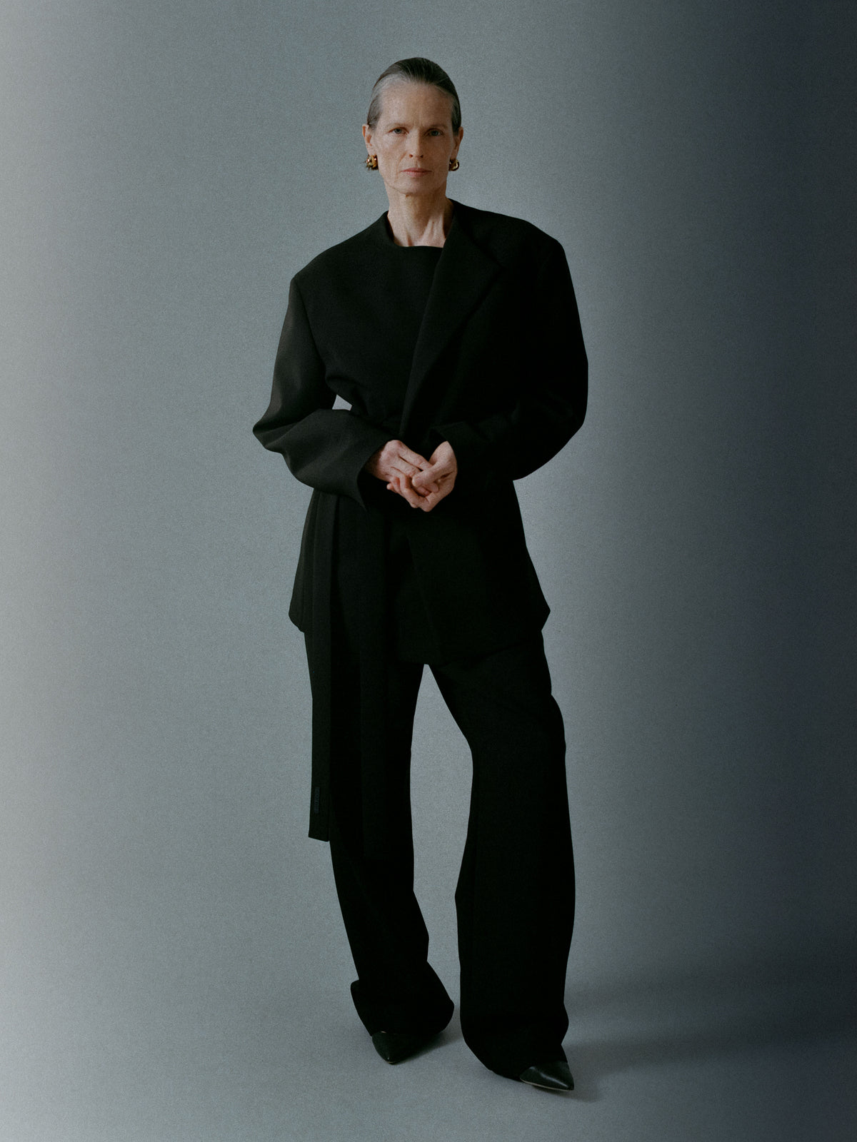 Woman wearing an oversized black blazer and wide-leg black pants standing against a minimalist gray background