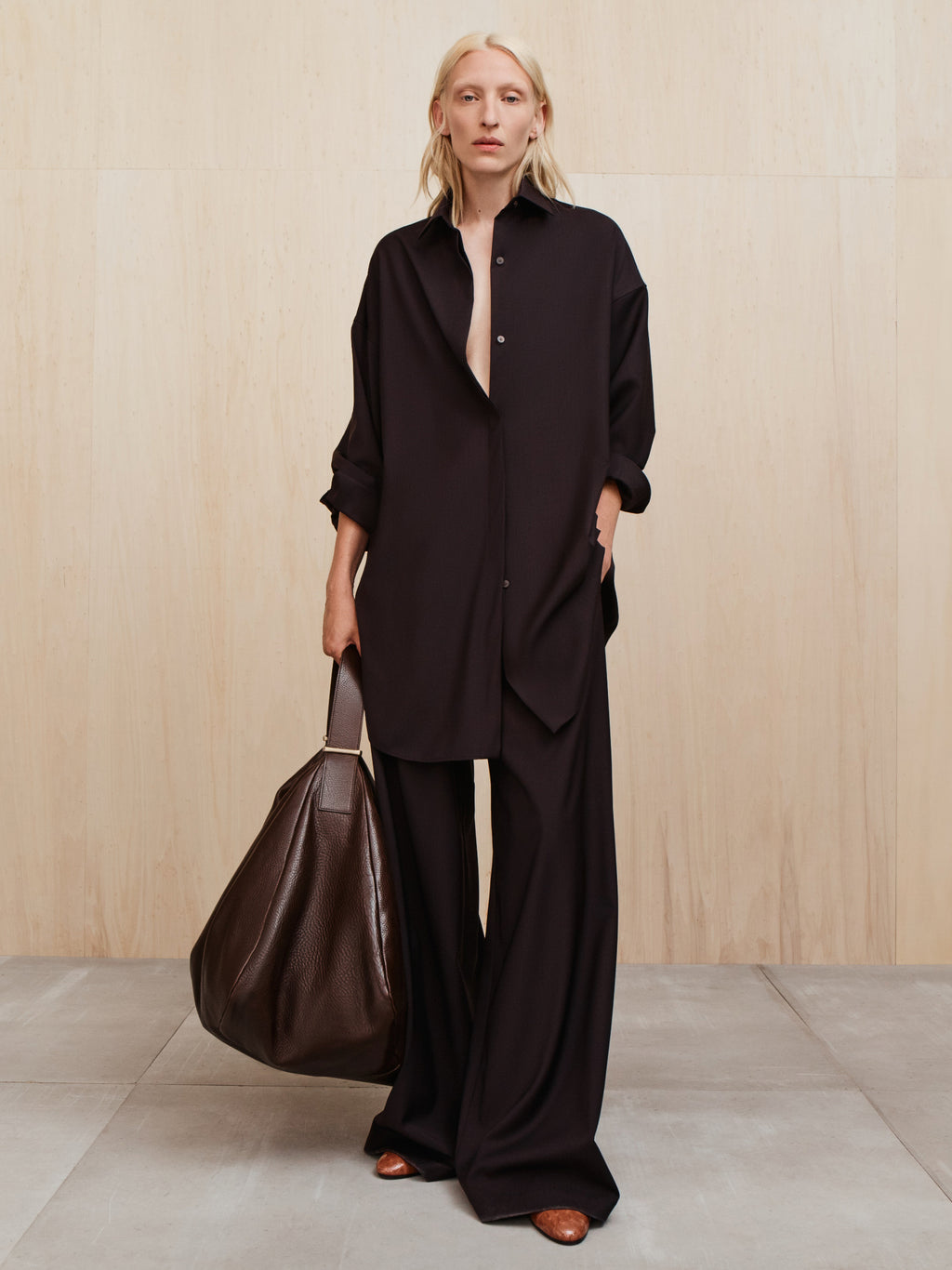 Person with light blonde hair wearing a loose black button-up shirt and wide black pants holding a large dark brown bag