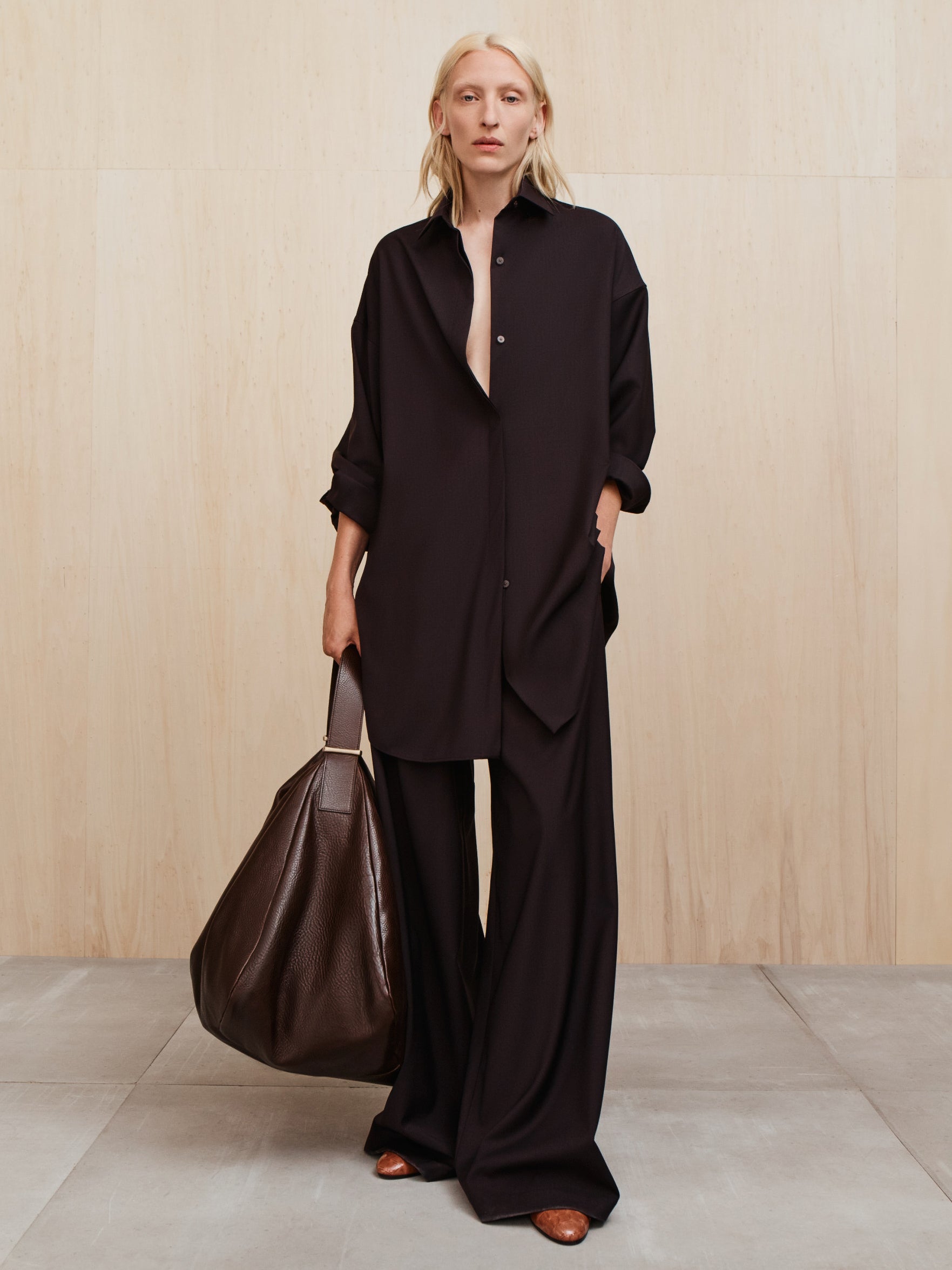 Person with light blonde hair wearing a loose black button-up shirt and wide black pants holding a large dark brown bag