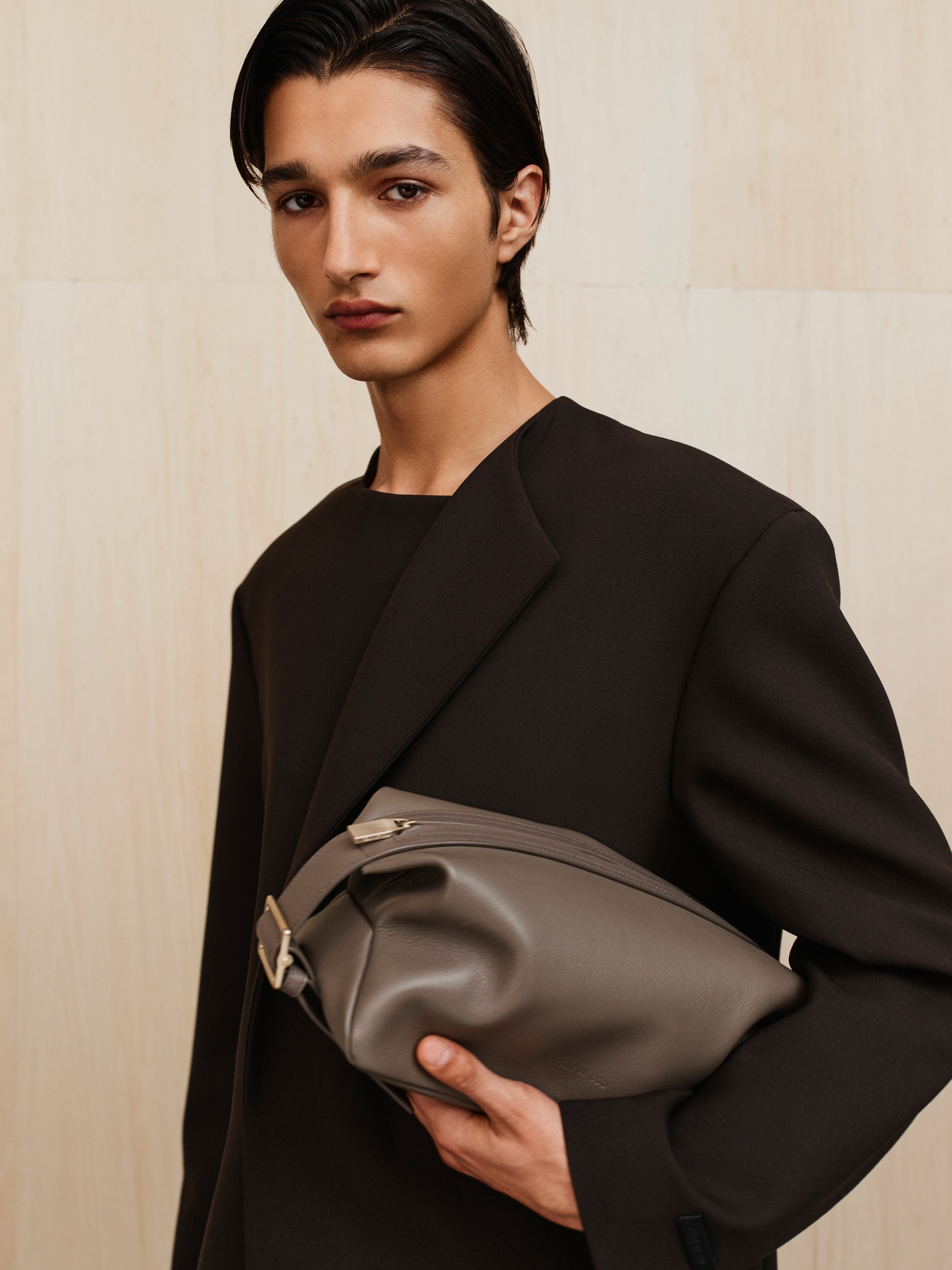 Young man in dark tailored jacket holding large gray leather pouch against beige wall background
